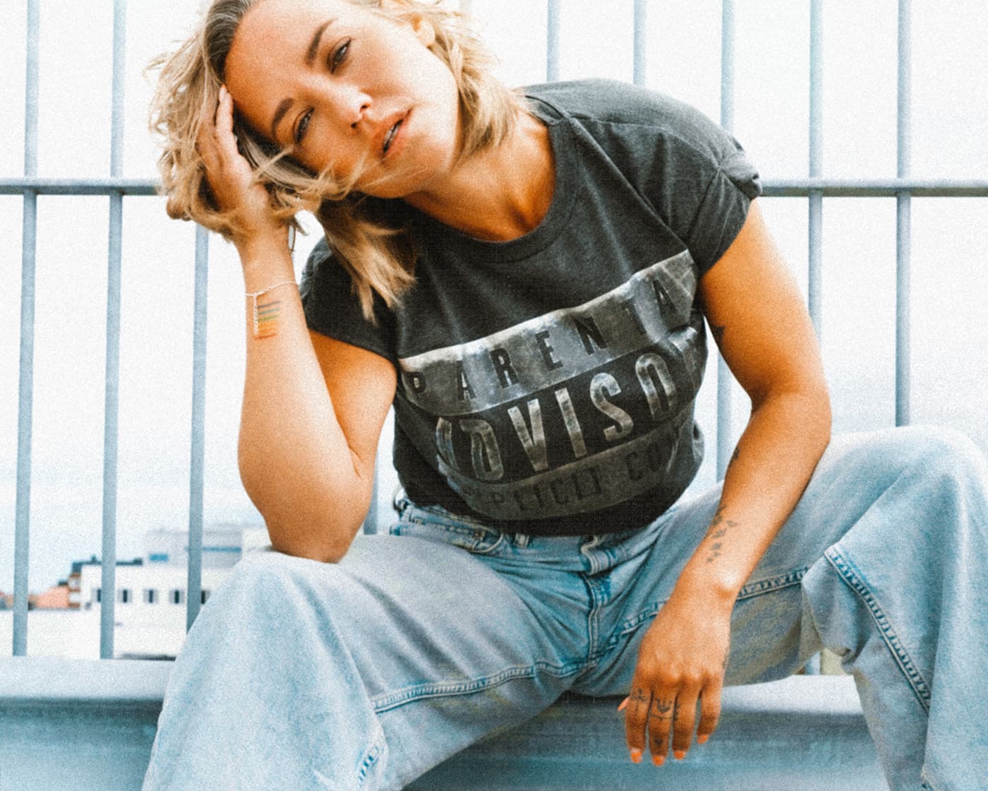 Color portrait of a blonde woman in a dark Harley-Davidson t-shirt and light jeans sitting in front of metal railings.