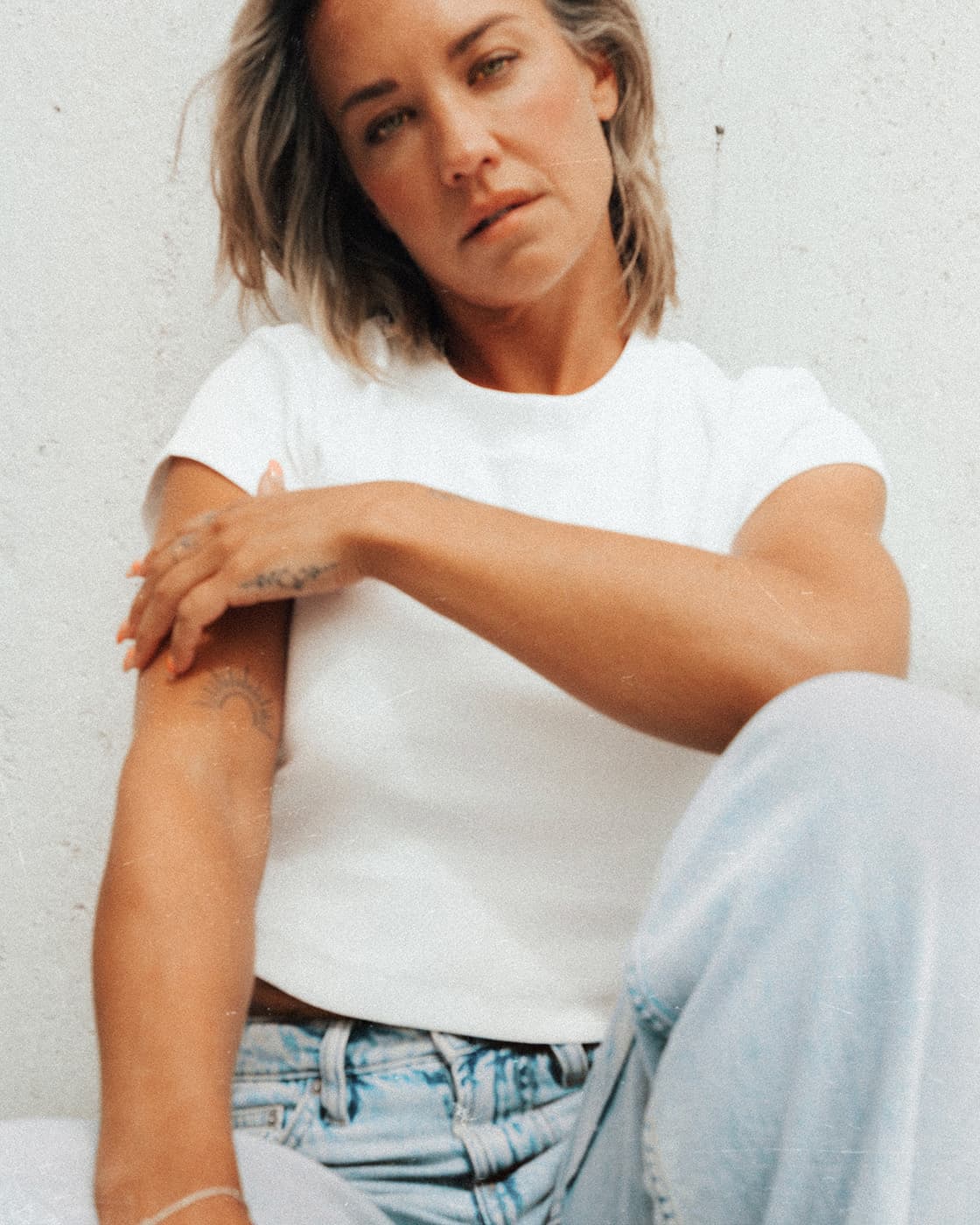 Color portrait of a woman in a white t-shirt and light jeans sitting against a pale wall.