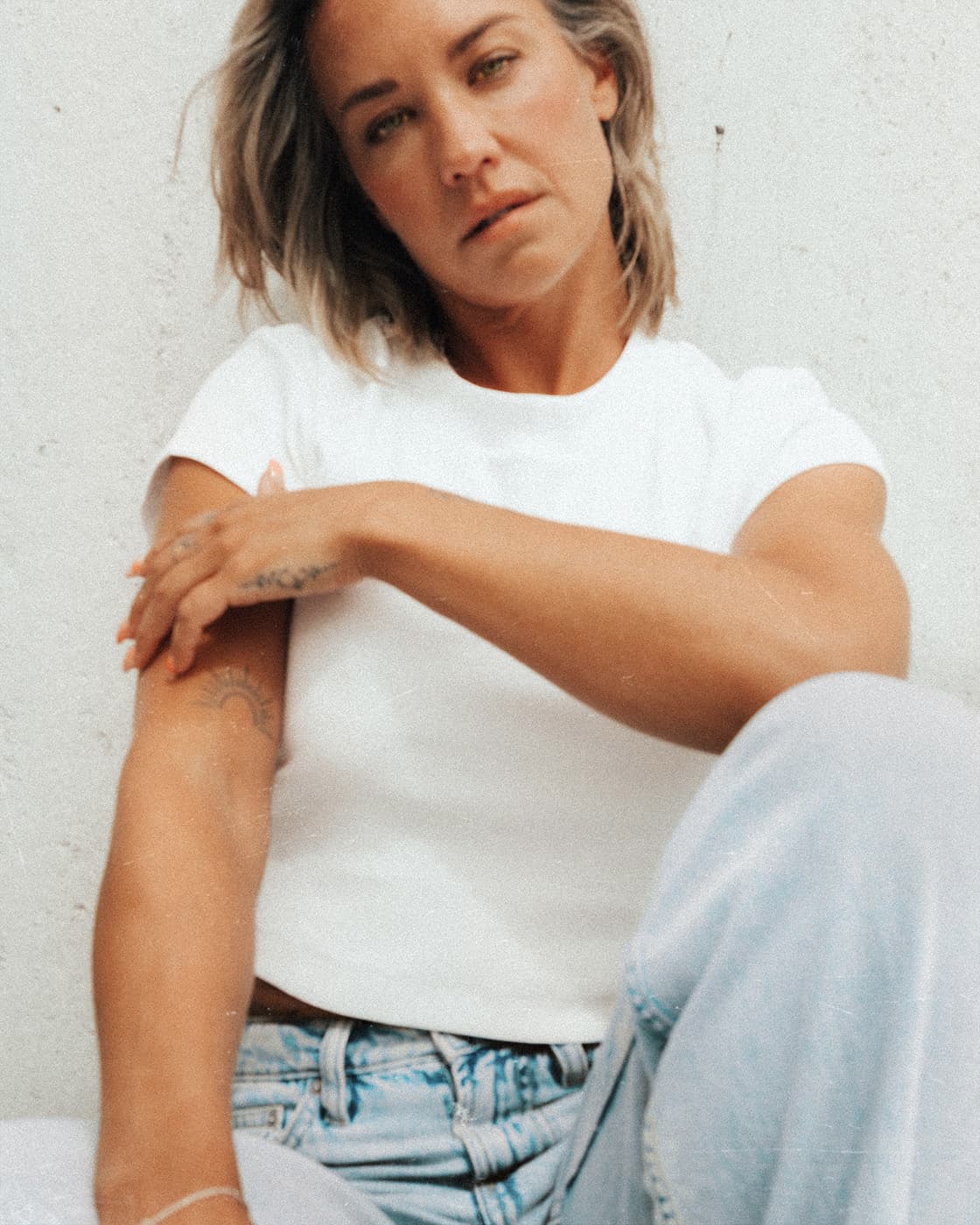 Color portrait of a woman in a white t-shirt and light jeans sitting against a pale wall.