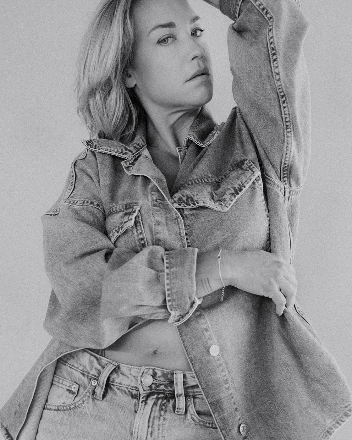 Black and white portrait of a blonde woman in a denim jacket with one arm raised.