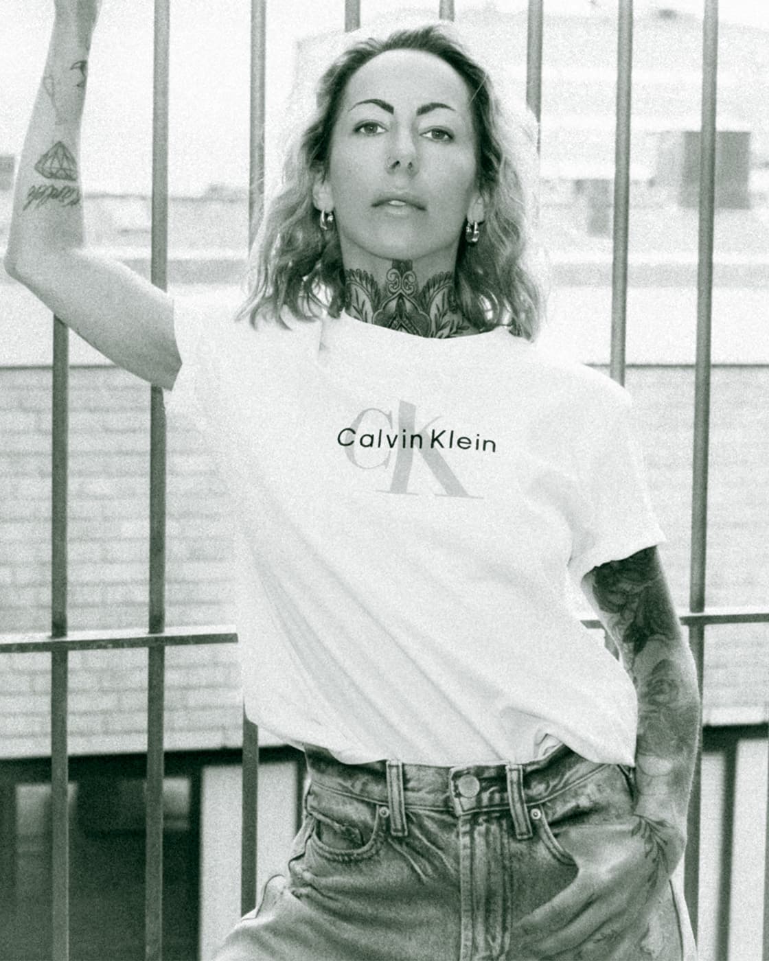 Black and white portrait of a tattooed woman wearing a white Calvin Klein t-shirt by metal bars.