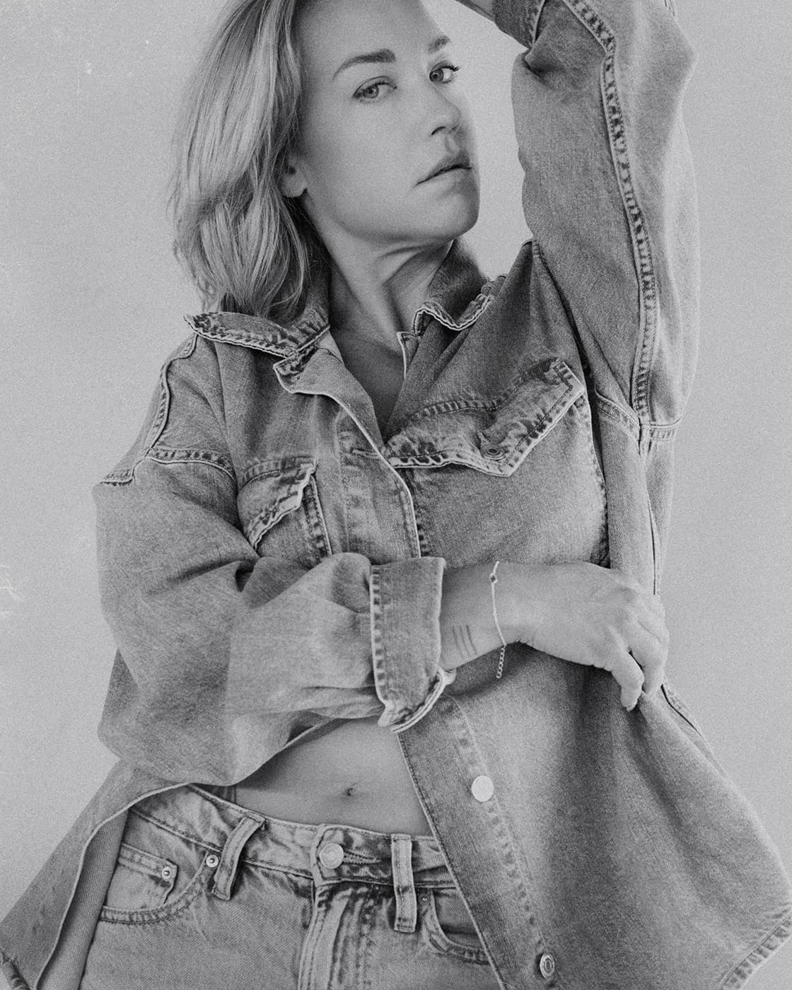 Black and white portrait of a blonde woman in a denim jacket with one arm raised.