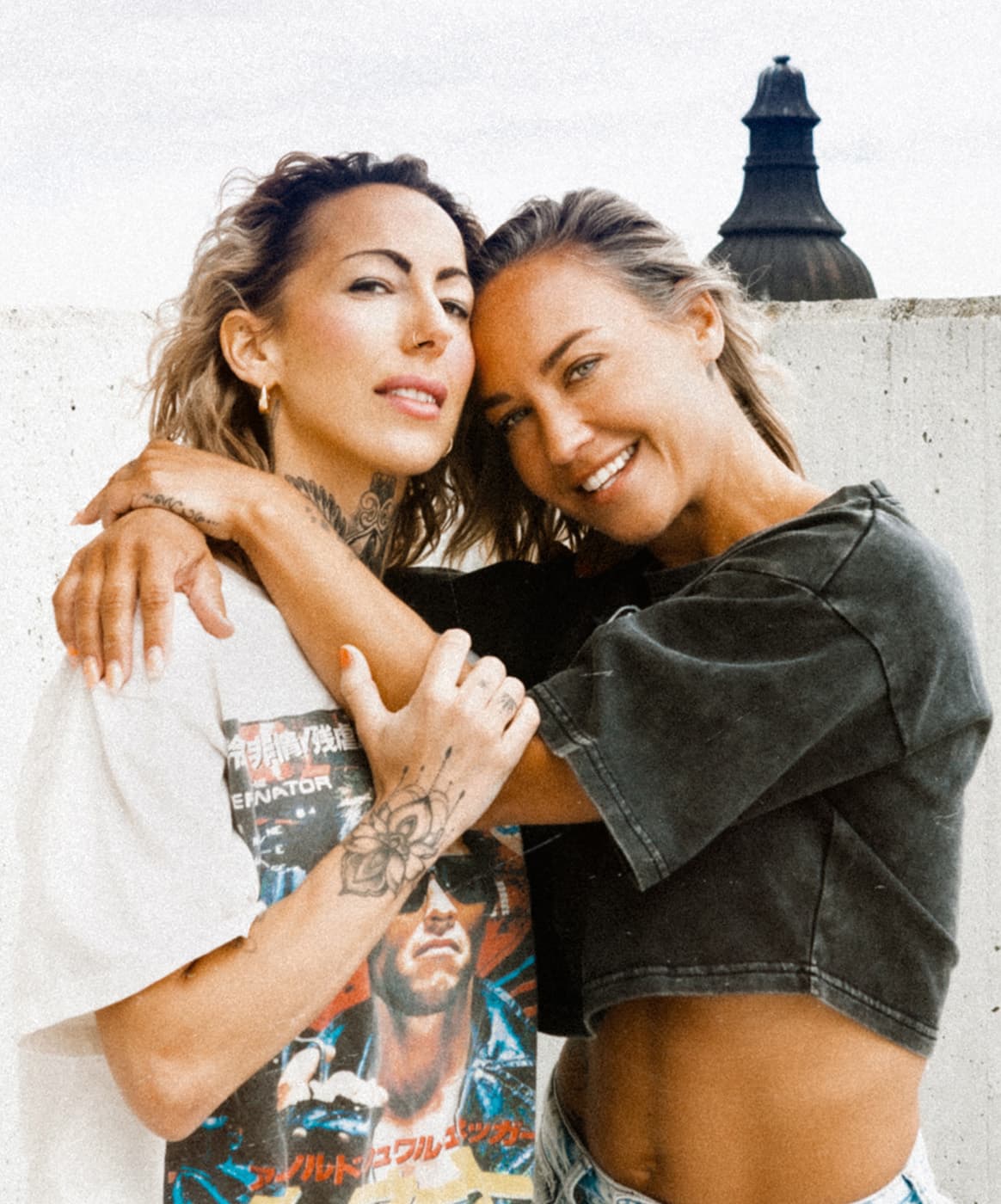Color portrait of two women standing close together in front of a pale wall.