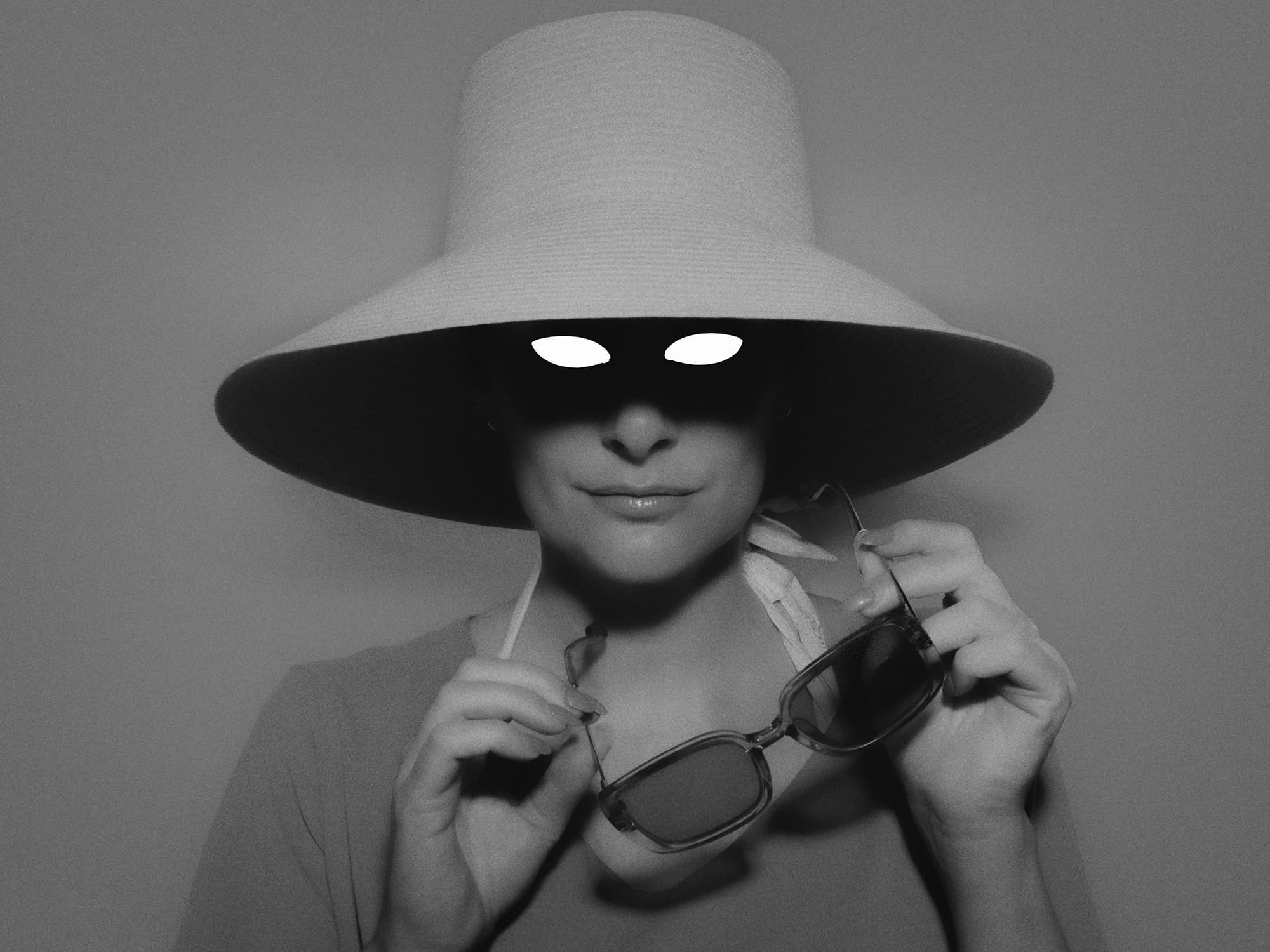 A portrait of a woman in a wide-brimmed hat holding sunglasses, with her eyes hidden in shadow.