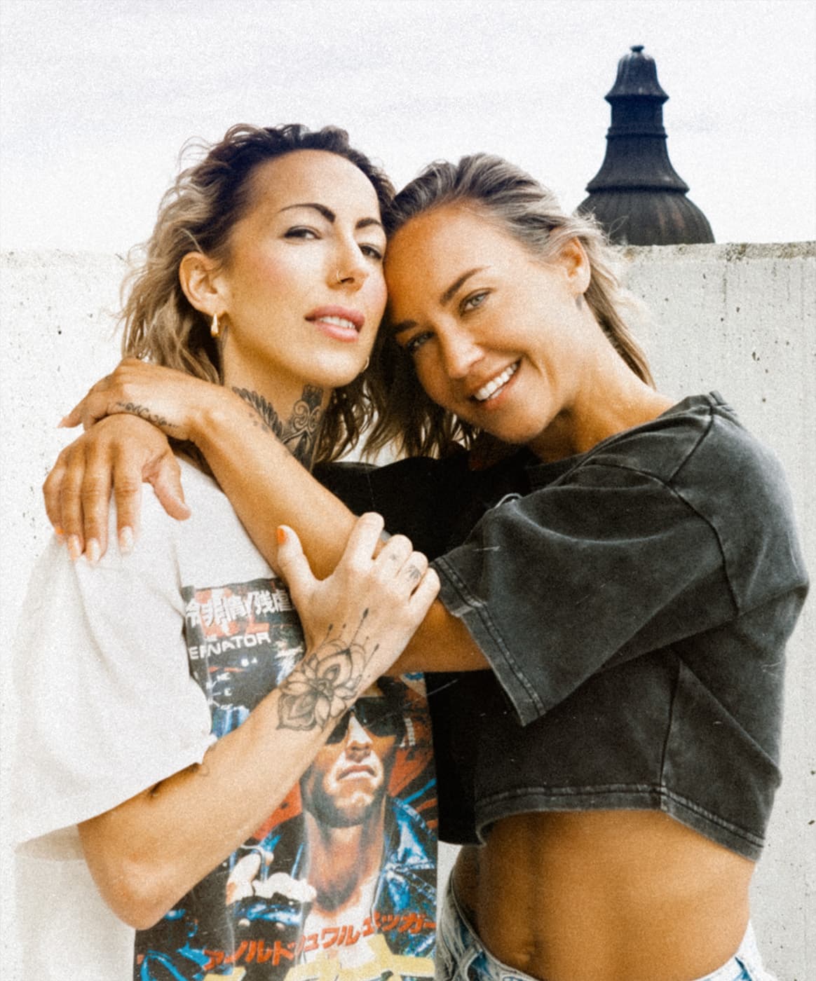 Color portrait of two women standing close together in front of a pale wall.