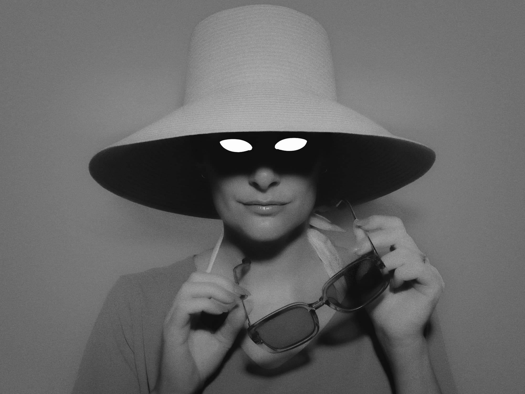 A portrait of a woman in a wide-brimmed hat holding sunglasses, with her eyes hidden in shadow.