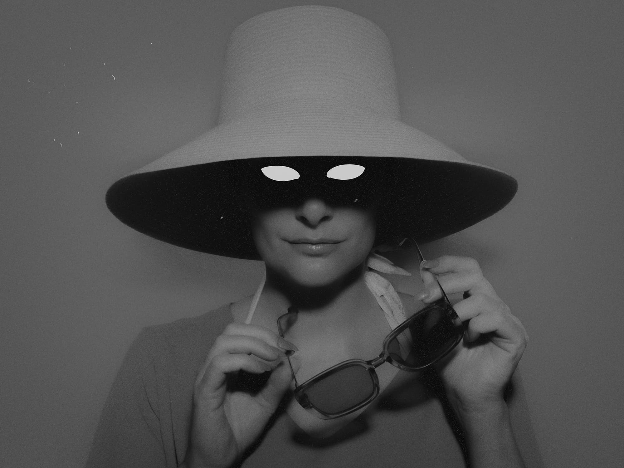 A portrait of a woman in a wide-brimmed hat holding sunglasses, with her eyes hidden in shadow.