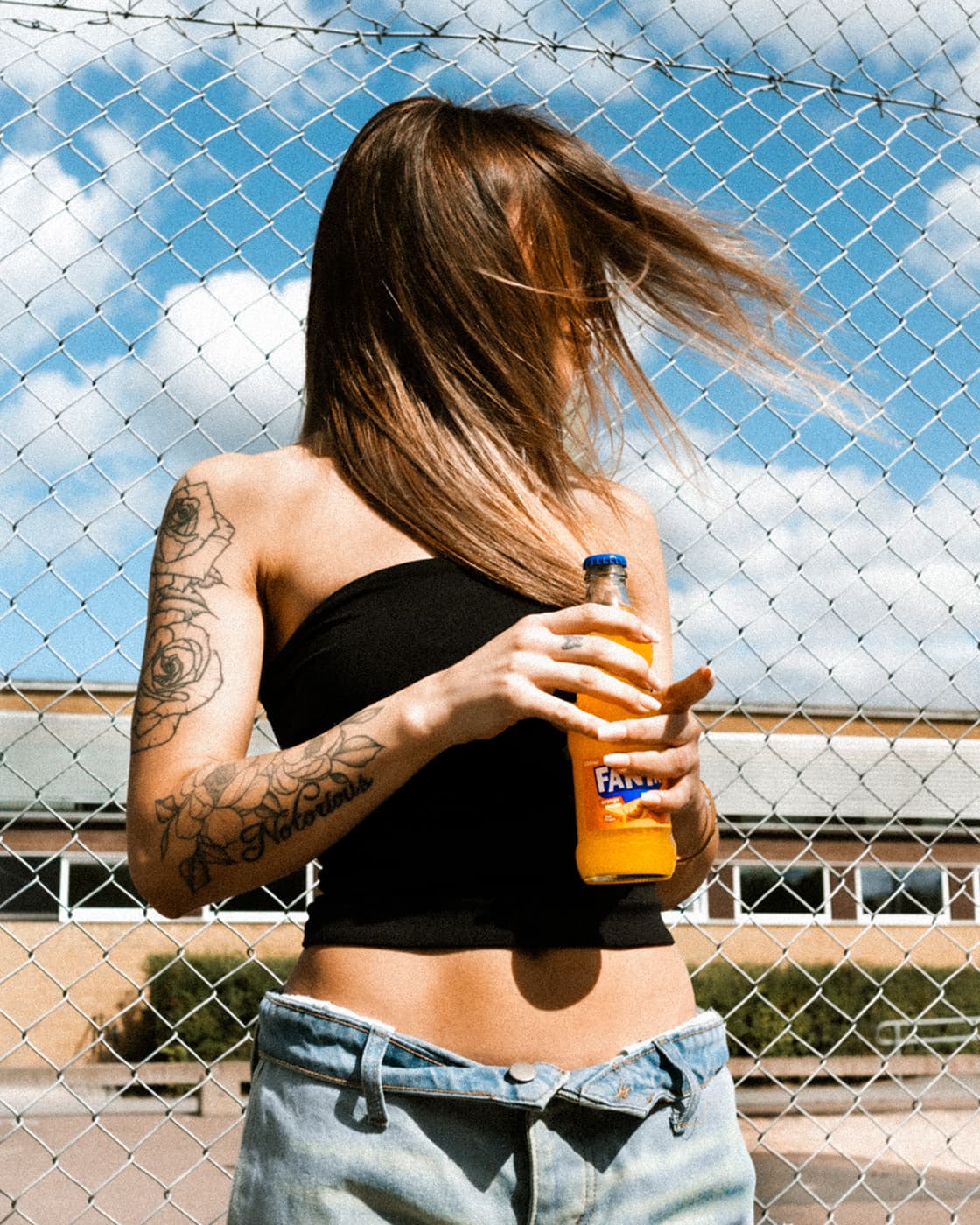 Color portrait of a woman holding an orange soda bottle in front of a chain-link fence.