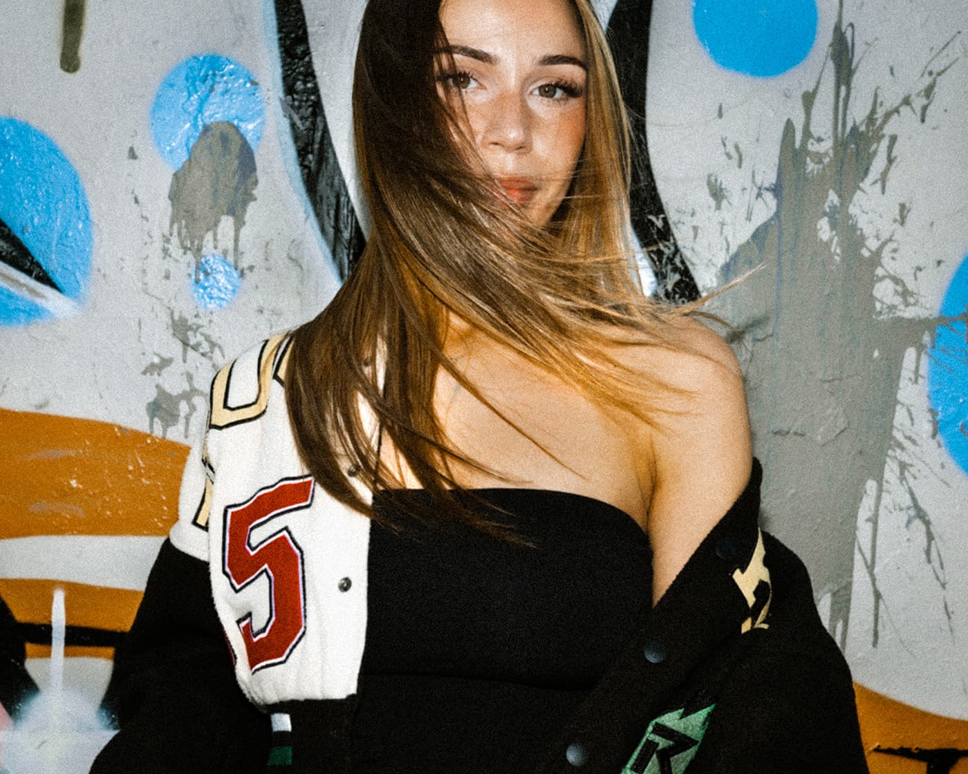 Color portrait of a woman with windblown hair wearing a black top and varsity jacket in front of a painted wall.