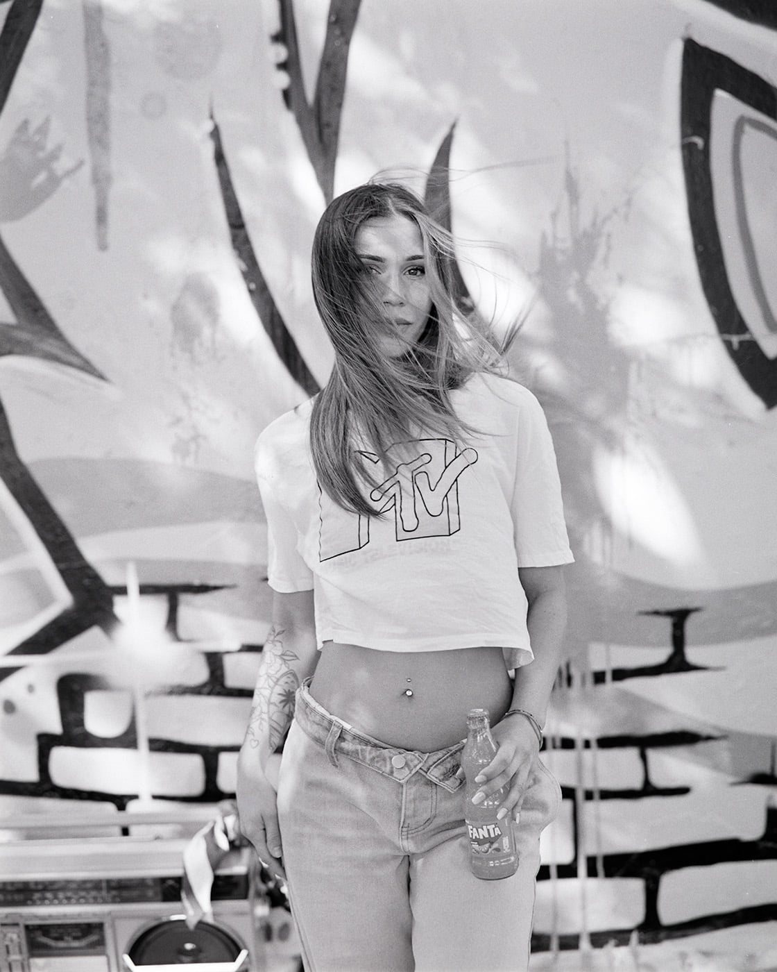 Black and white portrait of a woman in an MTV t-shirt holding a Fanta bottle in front of a graffiti wall.