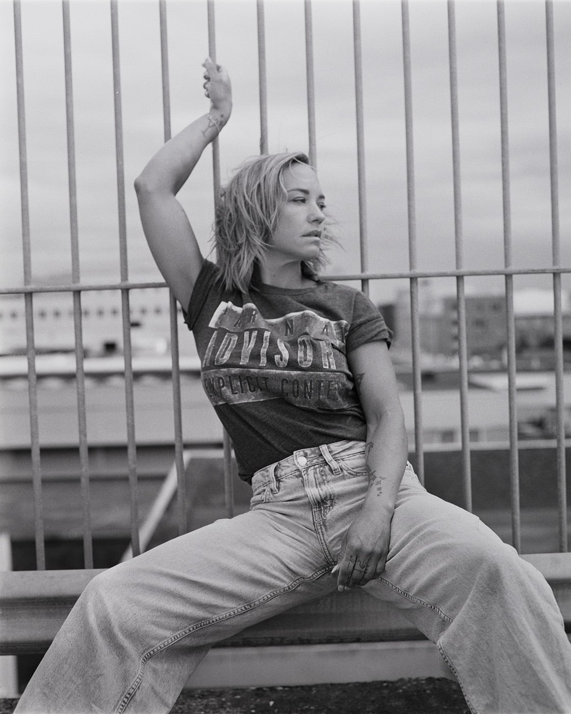 Black and white portrait of a blonde woman in a graphic t-shirt and light jeans sitting in front of metal railings with one arm raised.