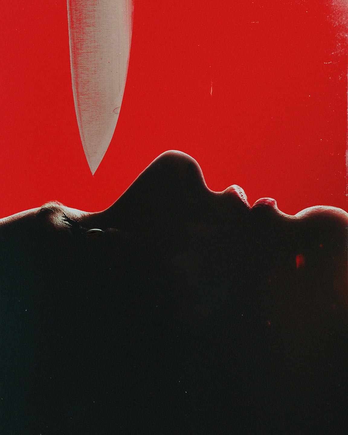Close-up of a face in shadow, seen in profile beneath a downward-pointing knife blade against a vivid red background.